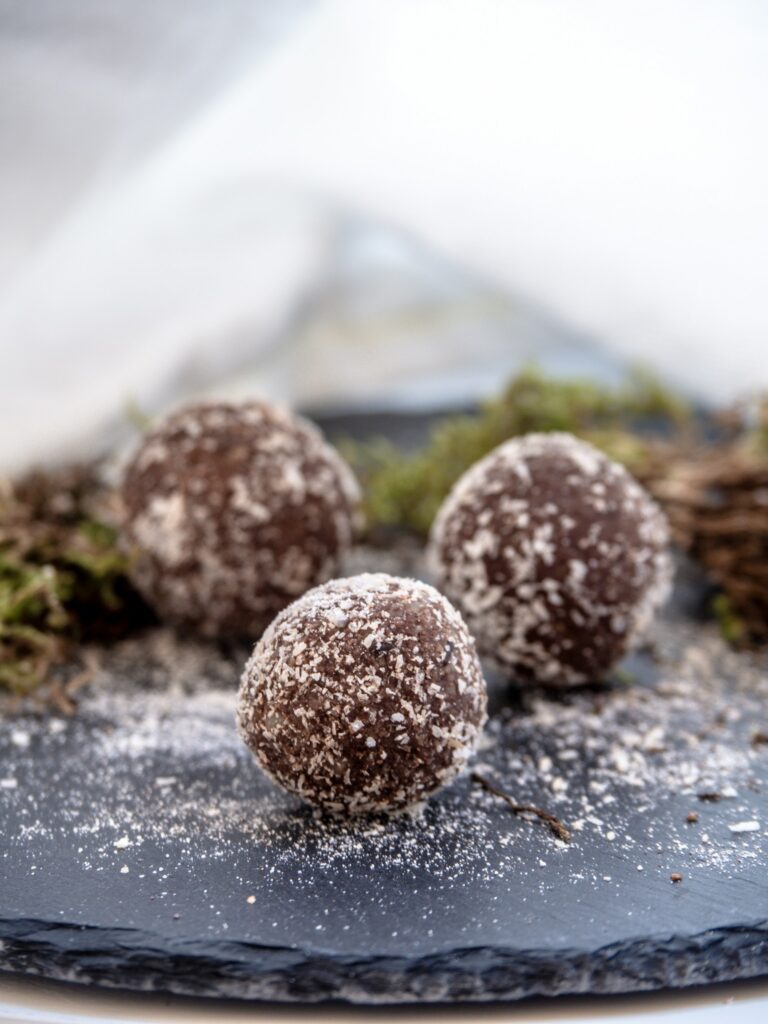 7 Hormone-Friendly Snacks Every Busy Mum Should Keep in Her Fridge | How to be a Mother y no Morir en el Intento chocolate and coconut pumpkin seed bliss balls on a dark plate, dusted with coconut, with a soft natural background.